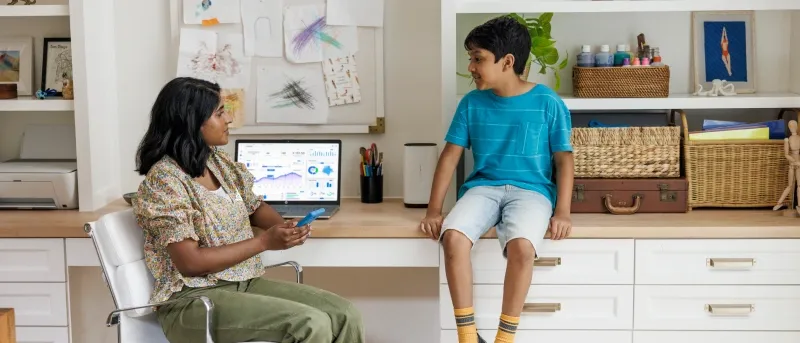 A mother sits in a white office chair looking at her son, who is sitting on a white and wood desk beside a laptop displaying charts and graphs.